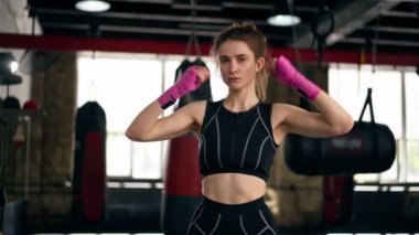 Bir boks kulübünde siyah kazaklı, pembe bandajlı genç bir kız antrenmana hazırlanmak için ısınma egzersizi yapıyor.