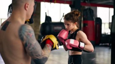 Bir boks kulübünde siyah kazaklı genç bir kız pembe eldivenli antrenörle yakın dövüş antrenmanı yapıyor.