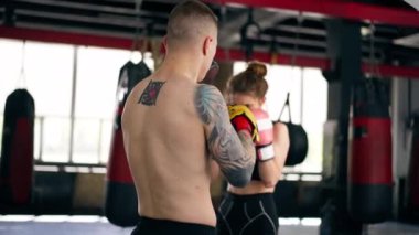 Bir boks kulübünde siyah kazaklı genç bir kız pembe eldivenli antrenörle yakın dövüş antrenmanı yapıyor.