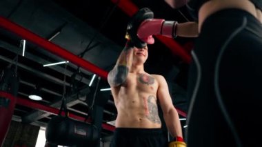 Bir boks kulübünde siyah kazaklı genç bir kız pembe eldivenli antrenörle yakın dövüş antrenmanı yapıyor.