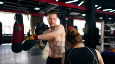 Bir boks kulübünde siyah kazaklı genç bir kız pembe eldivenli antrenörle yakın dövüş antrenmanı yapıyor.