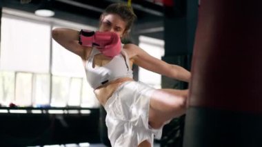 Bir boks kulübünde pembe eldivenli beyaz profesyonel üniformalı genç bir kız kırmızı bir çanta üzerinde teknik tekmeler atıyor.