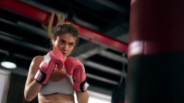 Dövüş kulübünde pembe eldivenli beyaz üniformalı genç bir kız kırmızı bir çanta üzerinde teknik tekme ve yumruk pratiği yapıyor.