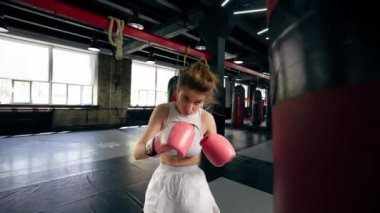 Dövüş kulübünde pembe eldivenli beyaz üniformalı genç bir kız kırmızı bir çanta üzerinde teknik tekme ve yumruk pratiği yapıyor.