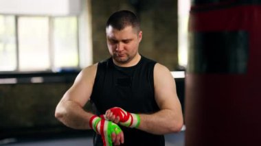 Bir boks kulübünde siyah tişörtlü yaşlı bir kas çalıştırıcı, kum torbasıyla çalışmadan önce ellerine çok renkli bir bandaj sarar.