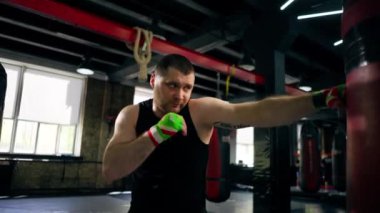 Bir boks kulübünde siyah tişörtlü yaşlı bir kas çalıştırıcı ve çok renkli bandajlarla boks yapıyor.