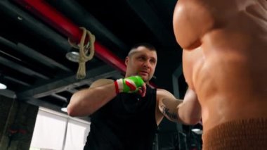Boks kulübü siyah tişörtlü yaşlı bir kas çalıştırıcısı ve çok renkli bandajlı bir insan mankeni boks yapıyor.