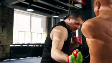 Boks kulübü siyah tişörtlü yaşlı bir kas çalıştırıcısı ve çok renkli bandajlı bir insan mankeni boks yapıyor.