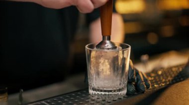Barın tezgahında güzel bir viski bardağı. Barmen buza pul yapıştırıyor.