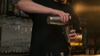 Önlüklü genç bir barmen alkol içeceği hazırlıyor. Kokteyl karıştırıcısına alkol dolduruyor.