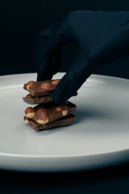 Siyah-mavi bir arka planda beyaz bir tabakta fındıklı süt çikolatası uzanıyor, parçalanmış bir el siyah bir eldiven seriyor.