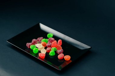 Siyah, uzun bir tabağa yakın. Çok renkli tatlı jöleler. Şekerli çeşitli meyveler serpiştirilmiş.