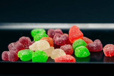 Siyah, uzun bir tabağa yakın. Çok renkli tatlı jöleler. Şekerli çeşitli meyveler serpiştirilmiş.