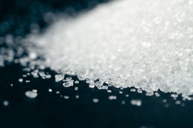 Siyah-mavi arka planda yakın plan beyaz granüle şeker uzanıyor, şeker yığınının etrafındaki kristal topaklar sağlığa zararlıdır