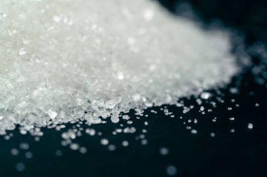 Siyah-mavi arka planda yakın plan beyaz granüle şeker uzanıyor, şeker yığınının etrafındaki kristal topaklar sağlığa zararlıdır