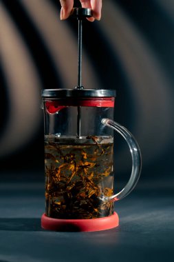 Koyu mavi arka planda, yan ışıklandırması olan bir cam modern çaydanlık var. Üzerinde yeşil çay demleyen bir el çabukluğu var.