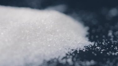 Siyah-mavi arka planda yakın plan beyaz granüle şeker uzanıyor, şeker yığınının etrafındaki kristal topaklar sağlığa zararlıdır