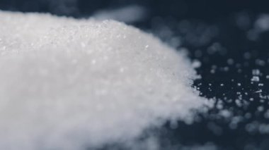 Siyah-mavi arka planda yakın plan beyaz granüle şeker uzanıyor, şeker yığınının etrafındaki kristal topaklar sağlığa zararlıdır