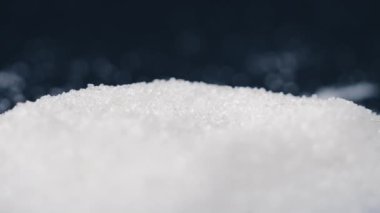 Siyah-mavi arka planda yakın plan beyaz granüle şeker uzanıyor, şeker yığınının etrafındaki kristal topaklar sağlığa zararlıdır