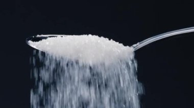 Siyah-mavi arka planda yakın plan beyaz granüle şeker uzanıyor üzerinde kristal şeker sağlığa zararlı bir kaşıktan dökülüyor