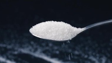 Siyah-mavi arka planda yakın plan beyaz granüle şeker uzanıyor üzerinde kristal şeker sağlığa zararlı bir kaşıktan dökülüyor