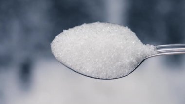 Siyah-mavi arka planda yakın plan beyaz granüle şeker uzanıyor üzerinde kristal şeker sağlığa zararlı bir kaşıktan dökülüyor