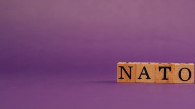 Siyah harfli, gölgeli tahta küplerle mor, ılık bir arka plan ve NATO harflerinden kelimeler.