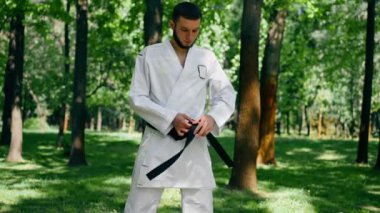 Parkta açık havada, güneşli havada genç karateci adam karate tekniğini kullanarak kemerini sıkıyor. Kara kuşak sağlıklı yaşam tarzı.