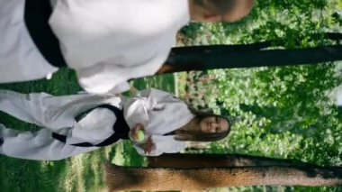 Güneşli havalarda parkta dikey video. Trenin çevikliği ve tepkisi olan genç bir karateci kız. Top dövüş sanatları kara kuşağıyla oynuyor.