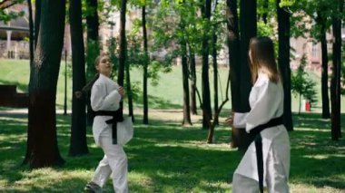 Parkta açık havada, güneşli havada genç bir karateci kız. Trenin çevikliği ve tepkisi. Siyah kuşakla oynayan bir top dövüş sanatları oyuncusuyla oynuyor.