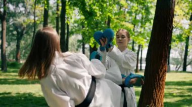 Parkta açık havada güneşli havada üniformalı iki karateci kız teknikleri tekmeliyor. Siyah kuşak dövüşü.