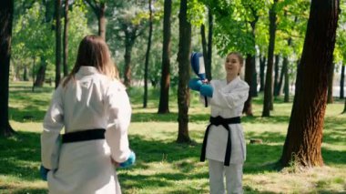 Parkta açık havada güneşli havada üniformalı iki karateci kız teknikleri tekmeliyor. Siyah kuşak dövüşü.