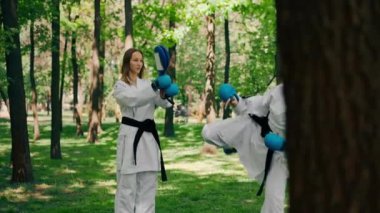 Parkta açık havada güneşli havada üniformalı iki karateci kız teknikleri tekmeliyor. Siyah kuşak dövüşü.