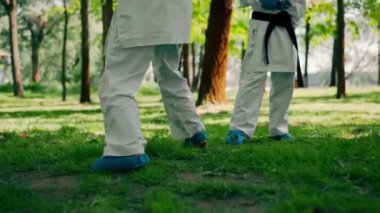 Parkta açık havada güneşli havada üniformalı iki karateci kız teknikleri tekmeliyor. Siyah kuşak dövüşü.