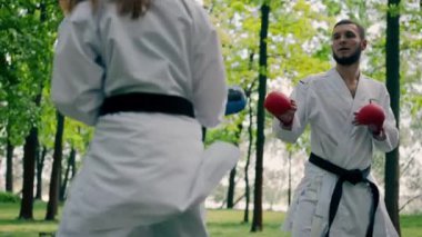 Parkta açık havada, güneşli havada üniformalı karateci kız teknikleri eğitir. Eşli siyah kuşaklı bir çift antreman maçı yapar.