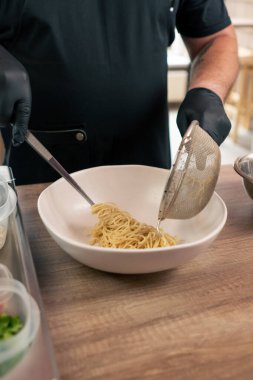 Bir fast food restoranında siyah üniformalı büyük bir şef önceden hazırlanmış malzemelerden Japon udon çorbası hazırlar ve beyaz tabağa erişte koyar.