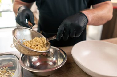 Bir fast food restoranında siyah üniformalı büyük bir şef önceden hazırlanmış malzemelerden Japon udon çorbası hazırlar ve beyaz tabağa erişte koyar.