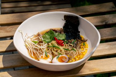 Sokaktaki bir restoranda, ahşap bir masanın üzerinde Japon udon çorbası, yumurta ve erişte karışımı beyaz bir tabak güneşte Japon hafif çorbası.