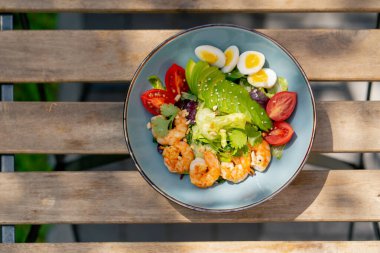 Caddedeki bir restoranda, bir kalas masanın üzerinde sıcak bir salata ve güneş ışınlarında karides olan mavi bir tabak.