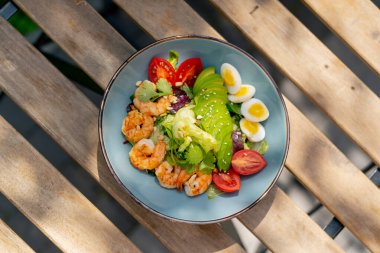 Caddedeki bir restoranda, bir kalas masanın üzerinde sıcak bir salata ve güneş ışınlarında karides olan mavi bir tabak.