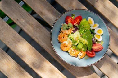 Caddedeki bir restoranda, bir kalas masanın üzerinde sıcak bir salata ve güneş ışınlarında karides olan mavi bir tabak.