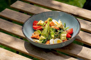 Üst çekim, caddedeki bir restoranda, bir tahta masanın üzerinde. Avokado ve güneş ışınlarında karides içeren sıcak bir salatayla mavi bir tabak.