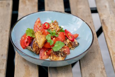 Yakın plan çekimde, sokağın ortasındaki bir lokantada, tahta masanın üzerinde. Sıcak salatalı, güneşte kızarmış tavuk ve domatesli mavi tabak.