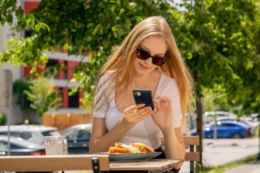 Caddedeki bir restoranda güneş gözlüklü güzel, uzun saçlı bir kız bir tabak yemeğin fotoğrafını çekiyor.