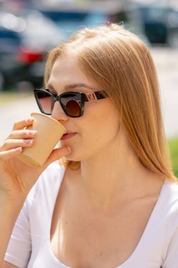 Caddedeki bir restoranda güneş gözlüklü güzel, uzun saçlı bir kız bir masada oturup eko bardaktan kahve içiyor.