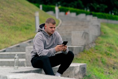 Parkta gri kazaklı genç bir adam taş bir merdivende oturmuş telefonda sohbet ediyor. Hava bulutlu.