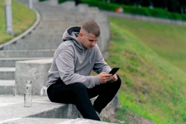 Parkta gri kazaklı genç bir adam taş bir merdivende oturmuş telefonda sohbet ediyor. Hava bulutlu.