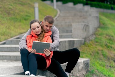 Parkta birbirine aşık genç bir çift spor yaptıktan sonra beton bir korkulukta sarılıyorlar, tablet bulutlu bir havada haberleri izliyorlar ortak bir hafta sonu.