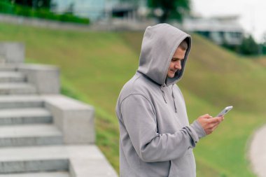 Parkta spor yaptıktan sonra gri kapüşonlu adam telefonda mesaj atıyor. Aktif, sağlıklı yaşam tarzı.