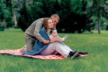 Parkta, kalın elbiseli bir adam ve kız, mavi bir battaniyeyle kaplı, kareli bir battaniyenin üzerinde kitap okuyan bir kıza sarılıyorlar.